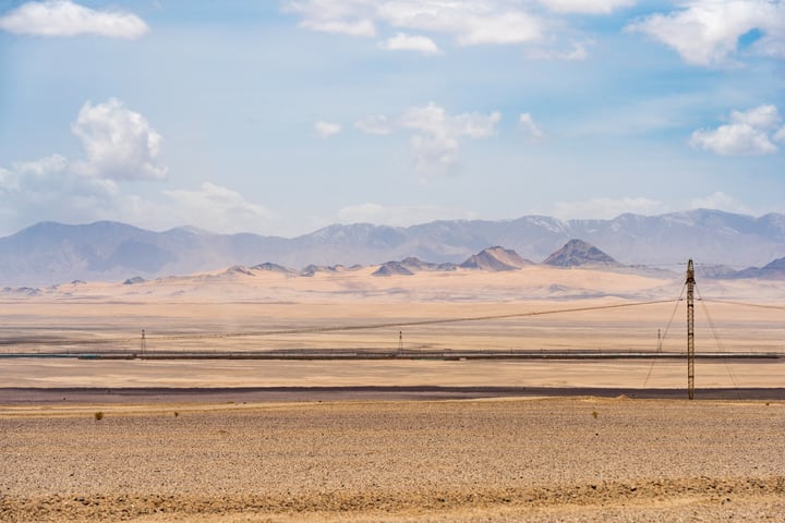 A vast desert landscape with a clear blue sky, mountains in the distance, and a few scattered clouds.