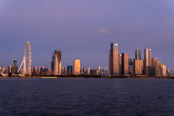 A serene cityscape is captured with a skyline of tall buildings, including a prominent Ferris wheel, against a backdrop of a calm body of water.