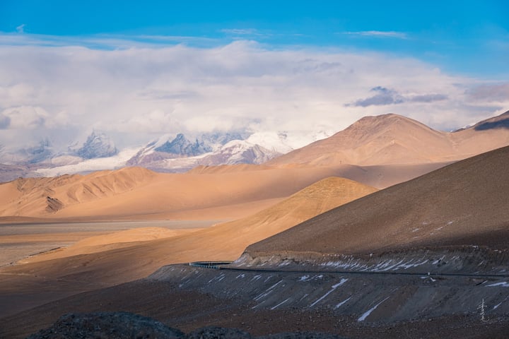A vast desert landscape with snow-capped mountains in the background, a blue sky with clouds, and a few scattered clouds.