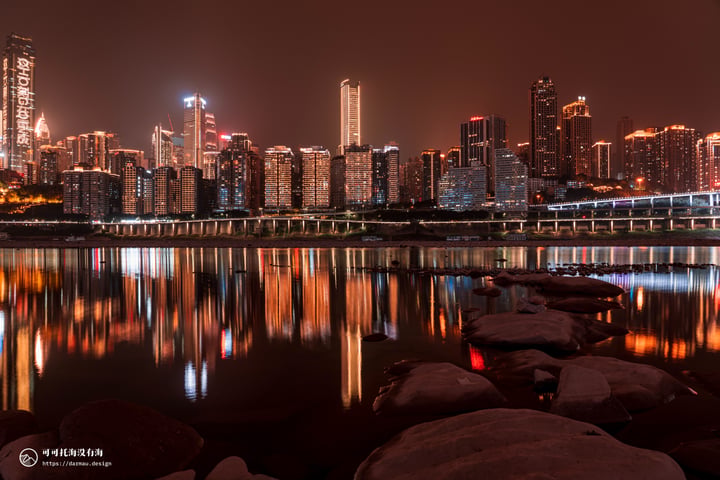 A nighttime cityscape is reflected in calm water, with tall buildings illuminated by lights and a bridge crossing the river.