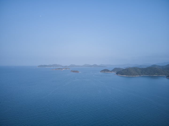 A clear blue sky with a few clouds, a calm ocean with small islands, and a small boat on the water.