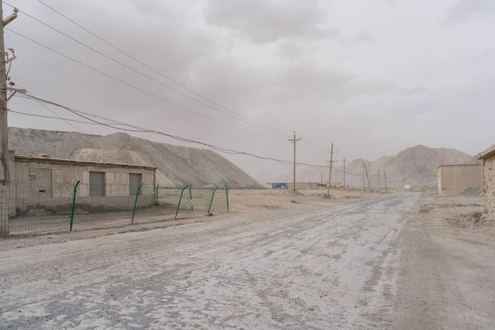 A desolate, barren landscape with a dirt road, a few buildings, and a mountain range in the background.