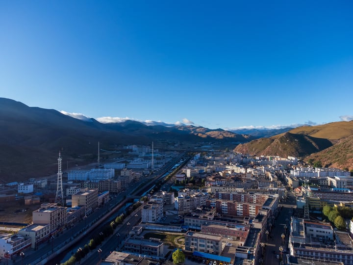 An aerial view of a city nestled between mountains, with a clear blue sky above, showcasing a mix of white and gray buildings, roads, and a river.