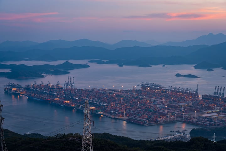 A large port with numerous cargo ships, cranes, and a large body of water with islands and mountains in the background.