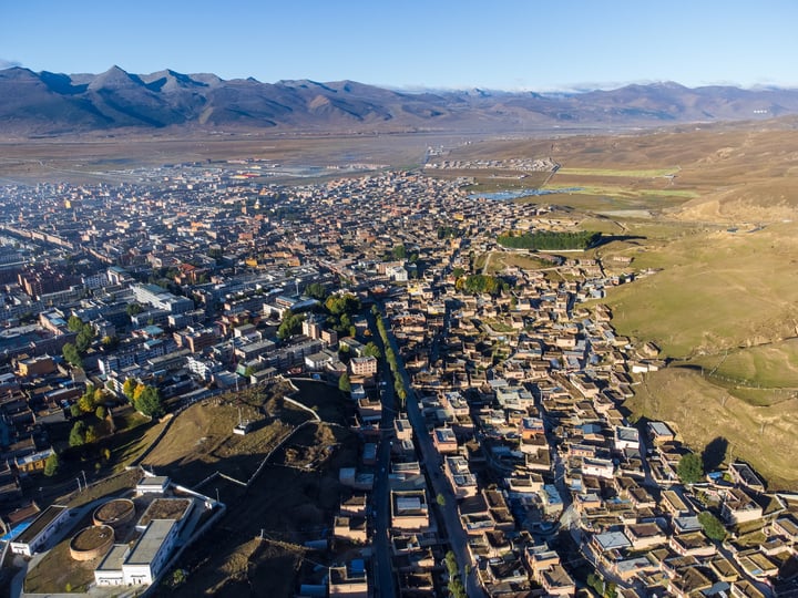 An aerial view of a city nestled between mountains, with a mix of buildings and green spaces, surrounded by snow-capped mountains.