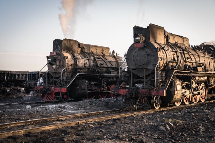 An old black steam locomotive with red wheels is traveling on a railway track, emitting smoke from its chimney.