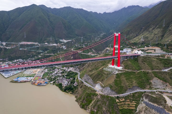 The image features the iconic suspension bridge in China, painted red and white, spanning a river. The bridge is supported by two white towers, and the surrounding landscape includes mountains and a town. The sky is cloudy, casting a soft light over the scene.