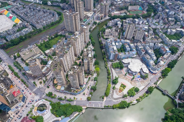 An aerial view of a city nestled between mountains, with tall buildings and a large white dome in the center, surrounded by lush greenery.