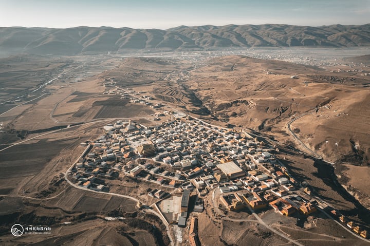 An aerial view of a town nestled in a valley, surrounded by mountains and surrounded by a vast expanse of green fields. The town is densely populated with white buildings, with a few orange and yellow structures scattered throughout. The sky is a clear blue, and the image is taken from above, providing a comprehensive view of the town and its surroundings.