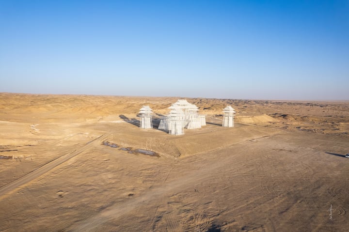 An aerial view of a white, pagoda-like structure with multiple levels and a central roof, surrounded by a barren desert landscape.