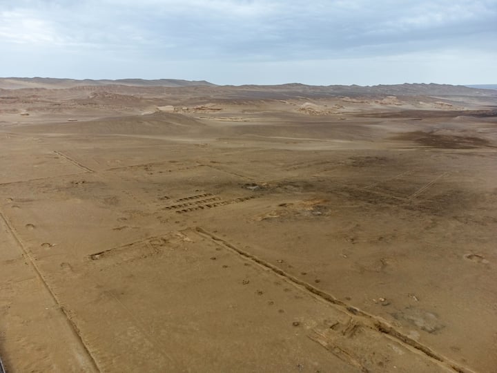 An aerial view of a vast desert landscape with sandy terrain, sparse vegetation, and a few small hills.