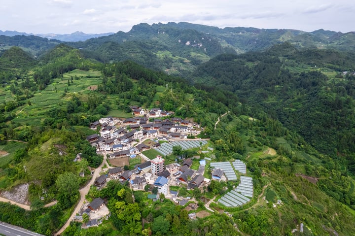 An aerial view of a small village nestled in a valley, surrounded by lush greenery and mountains, with a road running through the village and a road leading to the mountains.