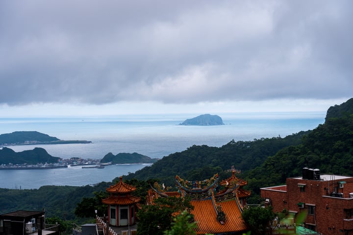 A serene coastal landscape features a large body of water with a few islands, a temple with a golden roof, and a mountain range in the distance.