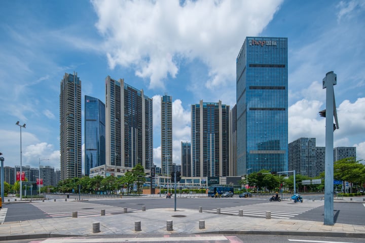 The image shows a cityscape with tall buildings, including skyscrapers, reflecting the blue sky with white clouds. The buildings are gray and blue, with the word "MALLA" written on them. In the foreground, there's a large square with a white line and a red sign. The sky is blue with white clouds.