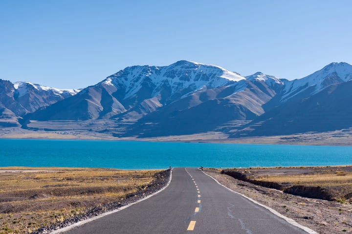 A serene landscape features a winding black road with a yellow center line, leading to a tranquil blue lake surrounded by snow-capped mountains.