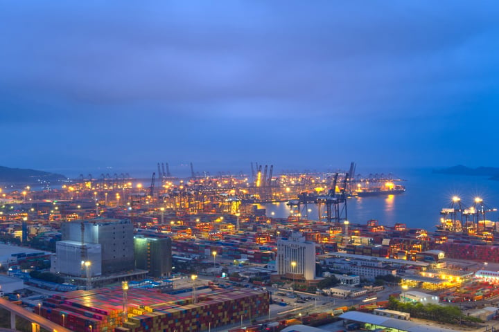 A cityscape at dusk, with a dark blue sky, illuminated cranes, and a harbor filled with colorful cargo ships.