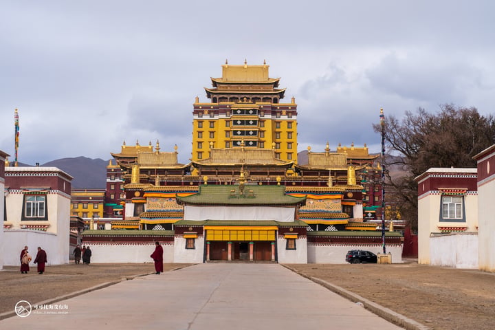 A grand, yellow temple with a red roof stands in a mountainous area, surrounded by a white fence and a few people walking around.