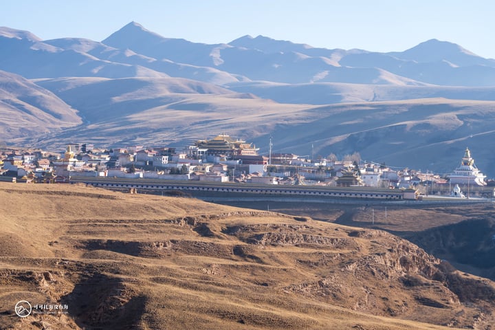 A serene mountain town nestled amidst brown hills, with white buildings and a golden dome, surrounded by majestic mountains under a clear blue sky.