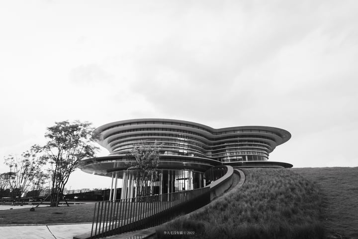 A modern, curved building with a large glass facade and a curved roof is surrounded by a grassy area and a fence.