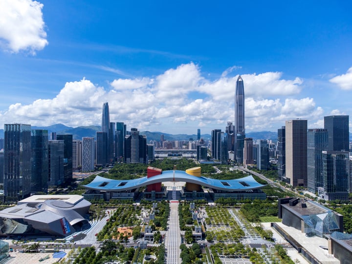 The image showcases a bustling cityscape with towering skyscrapers, a vibrant park, and a unique architectural feature. The park is surrounded by a large body of water, and the cityscape is framed by mountains in the background. The sky is a clear blue with a few clouds scattered across it.