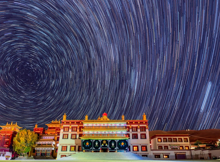 A grand, white-roofed temple with a red roof stands in a mountainous landscape, bathed in a starry sky. The temple's intricate architecture and vibrant colors contrast with the dark sky, creating a captivating scene.