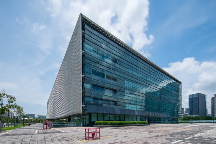 A modern, rectangular glass building with a curved facade stands in a bustling cityscape. The building's large windows reflect the city's life, and a red bench is positioned in front for visitors to rest. The sky is a clear blue with white clouds, and the city skyline is visible in the distance.
