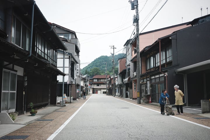 A woman pushes a cart on a street lined with buildings, with a hill in the background.