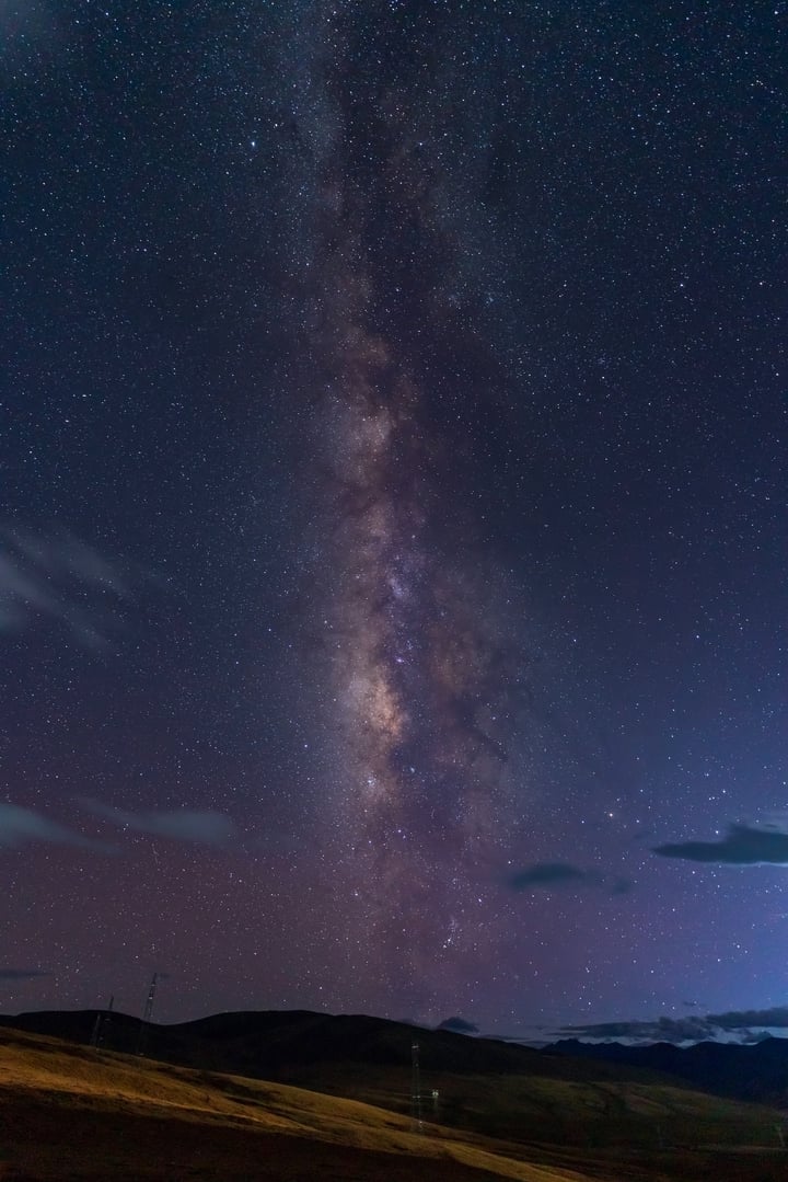 A night sky filled with stars and the Milky Way stretching across the sky. A large field with grass and hills in the background.