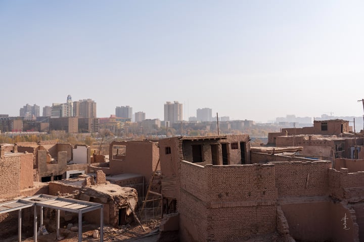 A cityscape with a mix of old and new buildings, including a red brick structure with a white roof, surrounded by a hazy blue sky and a distant city skyline.