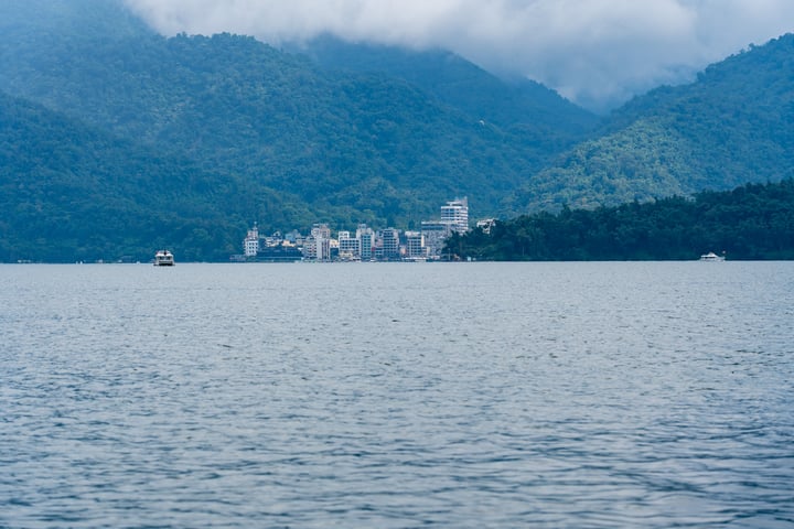 A serene mountainous landscape with a large body of water in the foreground, with boats scattered across the water, and a city in the background.