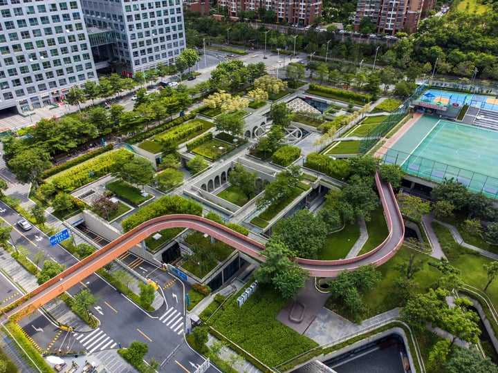 An aerial view of a city park with a curved red road, surrounded by lush greenery and a tennis court, with modern buildings in the background.