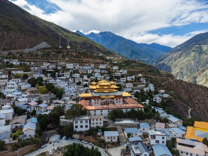 The image captures a serene mountain town nestled amidst lush greenery, with white buildings and a prominent golden temple. The temple, with its golden roof, stands out against the backdrop of towering mountains. The sky above is a clear blue, with scattered clouds adding depth to the scene. The town appears to be a small village, with a few cars parked nearby. The image is taken from a high vantage point, offering a panoramic view of the town and its surroundings.