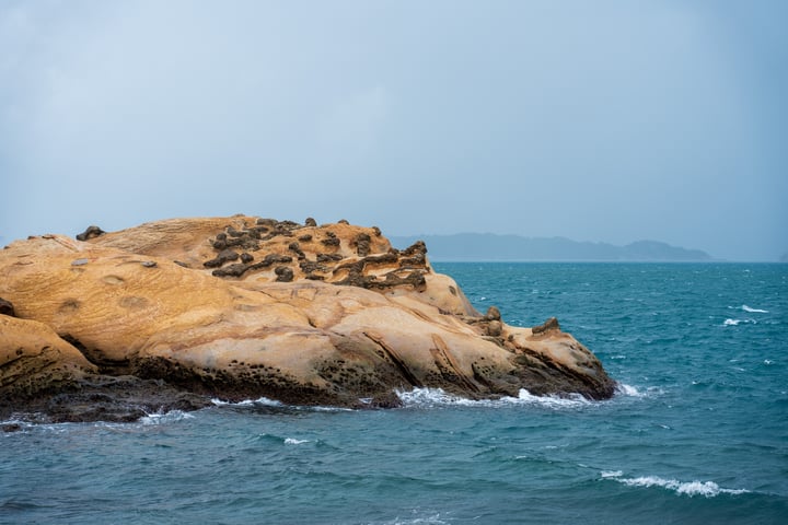 A rocky island with a few small structures, surrounded by deep blue water and a hazy sky, with a mountain range in the distance.