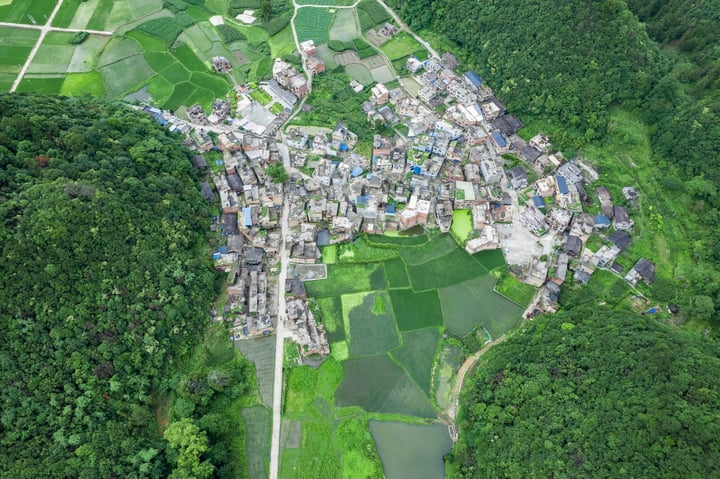 An aerial view of a village nestled amidst lush greenery, with a mix of traditional and modern buildings, surrounded by fields and water bodies.