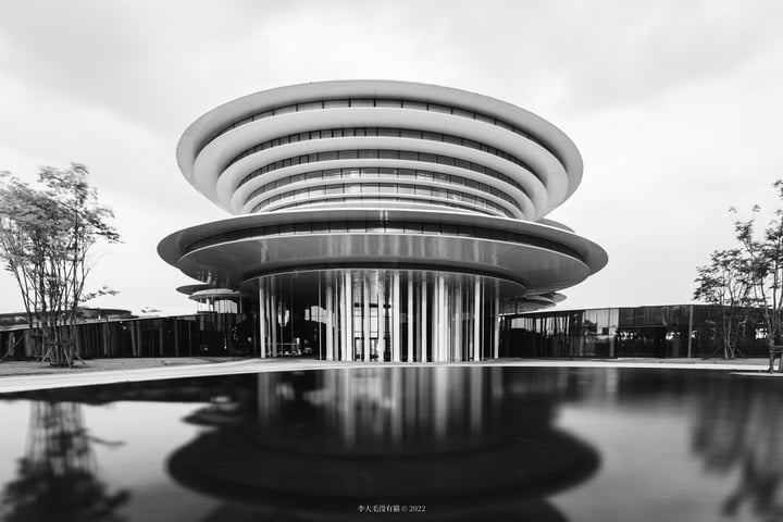 A modern, circular building with a curved roof and large windows is reflected in a pool, surrounded by trees and a cloudy sky.
