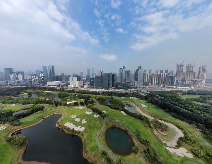 The image captures a serene golf course nestled amidst a bustling cityscape, with a clear blue sky overhead. The greenery of the course contrasts beautifully with the urban backdrop, while the city skyline in the distance adds a dynamic element to the scene.