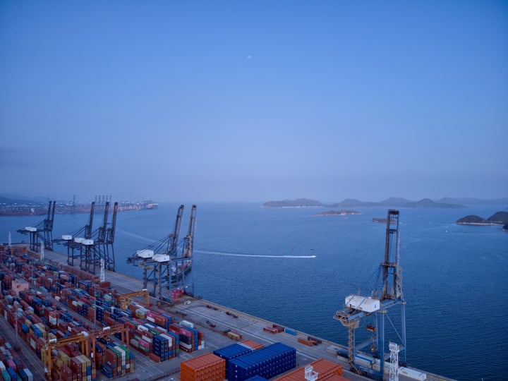 A bustling harbor scene with a large cargo ship, numerous boats, and cranes, with a clear blue sky and a small moon in the distance.