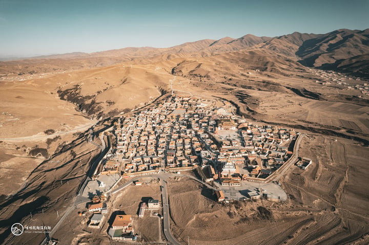 An aerial view of a desert town with a mix of white and brown buildings, surrounded by mountains and a road, showcasing a blend of nature and human habitation.