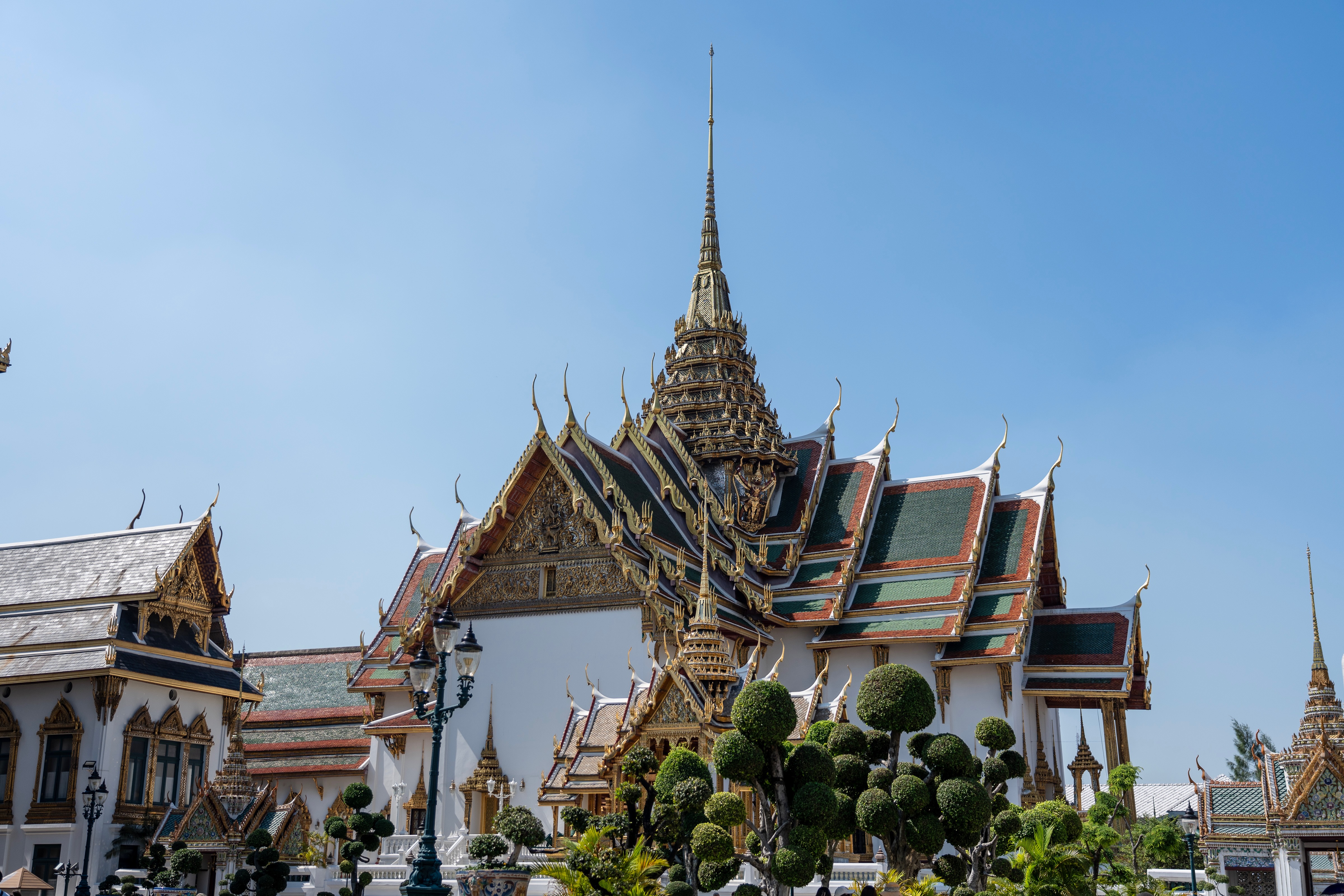 The image showcases the iconic Wat Arunath, a temple complex in Bangkok, Thailand. The temple complex features ornate gold and white structures, with a central spire reaching towards the sky. The image is taken from a low angle, emphasizing the grandeur of the temple and the surrounding greenery.