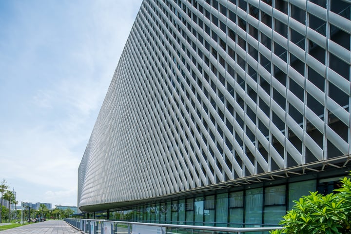 A modern building with a geometric facade and a grid-like pattern, featuring large windows and a metal railing, stands tall against a clear blue sky. The building is situated on a street with trees and lampposts in the foreground, creating a harmonious blend of architecture and nature.