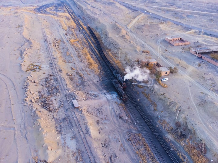 An aerial view of a black train engine emitting steam on a track, surrounded by a barren landscape with a red brick building and a white truck.
