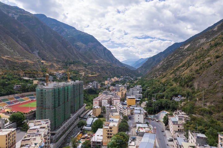 A city in a valley with mountains, surrounded by construction cranes, is captured in a high-angle view. The city has a mix of residential and commercial buildings, with a soccer field in the foreground. The sky is filled with clouds, and the mountains are covered in green vegetation.