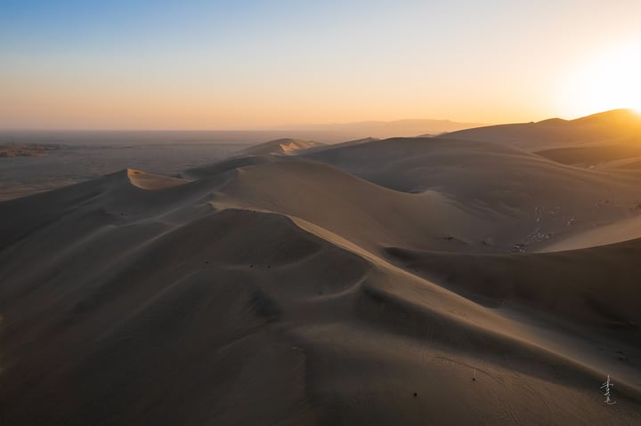 A serene desert landscape at sunset, with a large sand dune in the foreground and smaller dunes in the background. The sun is setting, casting a warm glow on the sand and creating a tranquil atmosphere.