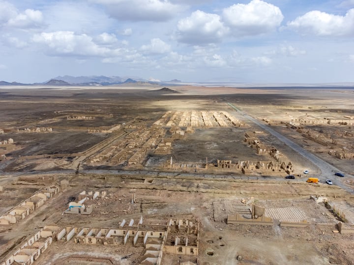 An aerial view of a desert landscape with a large ancient city in the center, surrounded by a vast expanse of dirt and mountains, with a road running through it.