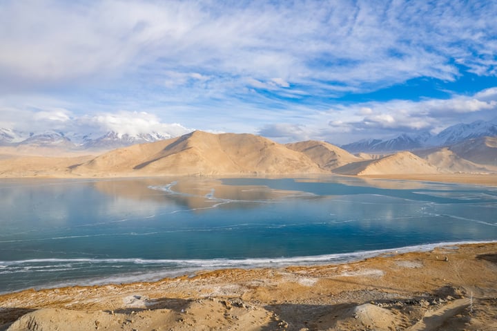 A serene landscape features a deep blue lake reflecting the sky, with snow-capped mountains in the distance. The sky is filled with white clouds, and the landscape is dominated by brown and beige tones.
