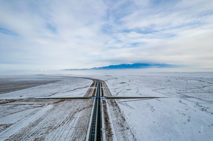 An aerial view of a snow-covered road leading to mountains, with a clear blue sky above.