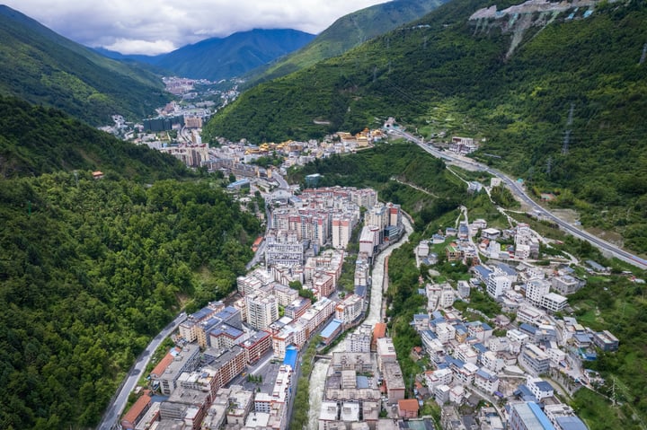 An aerial view of a mountain town with a winding road, surrounded by lush greenery and towering mountains, showcasing a harmonious blend of nature and human habitation.