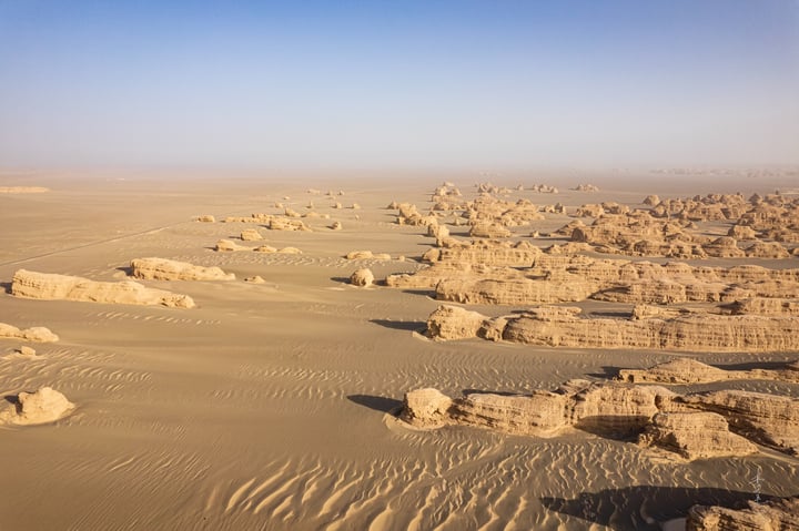 Aerial view of a desert landscape with sandy formations, a clear blue sky, and a few distant structures.