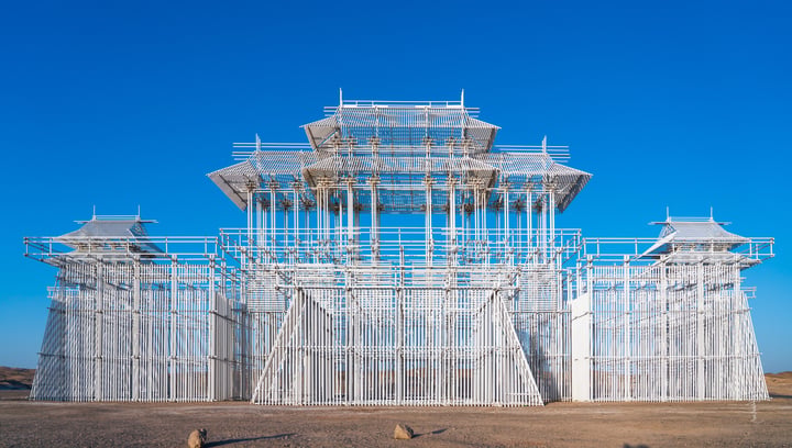 A large, white, glass-enclosed structure with multiple levels stands in a desert-like environment, surrounded by a clear blue sky.