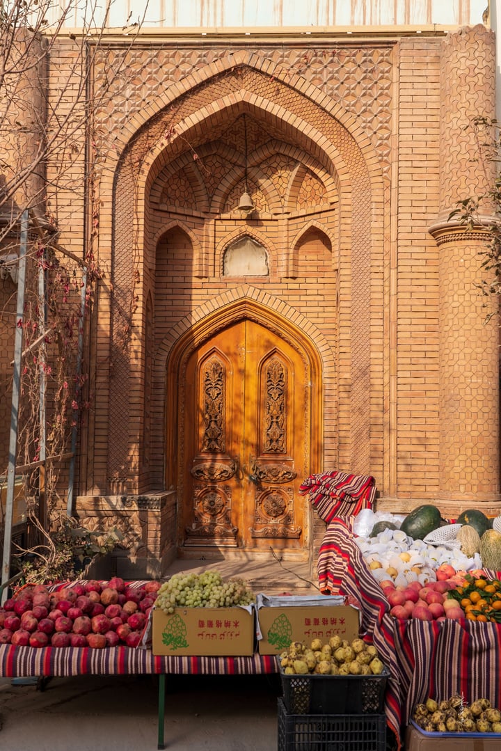 A narrow cobblestone street in a traditional Kashgar is lined with pink and brown buildings, some with green and red accents. An archway with green and red patterns leads to a red door, and potted plants are scattered around. The sky is clear, and the sun shines